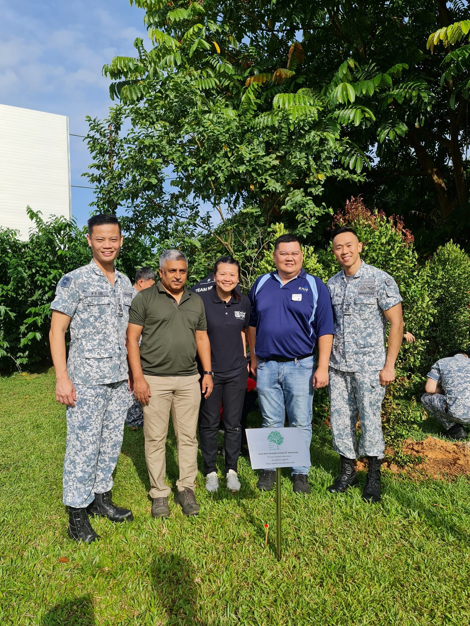 Five people pose outdoors near a small planted tree with a commemorative sign, two wearing military camouflage uniforms.